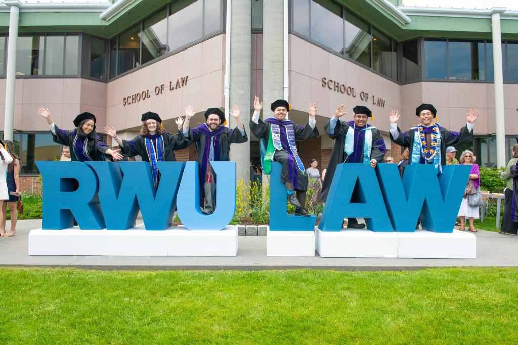 Graduating law students wearing cap and gown posing by large RWU LAW letters in front of the Law School building.