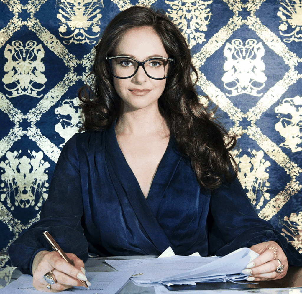 Carrie goldberg, Woman wearing glasses seated at a desk, holding a pen and papers, against a blue patterned background