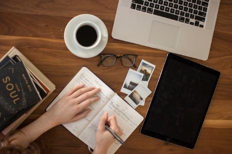 Books, laptop, eye glasses, hand of person writing in a notebook, and a cup of coffee