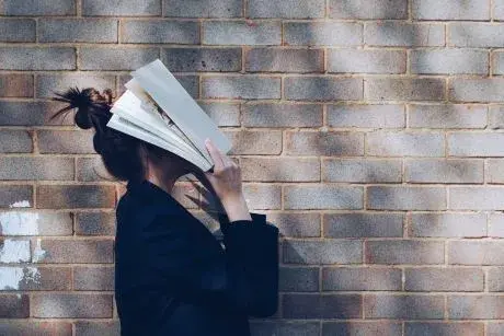 Person with bun on top of head  wearing a black shirt standing in front of brick wall and holding an open book over their face.