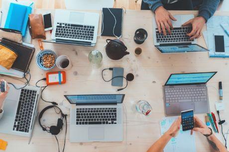 Table top with several laptop computers, phones, tablets, and coffee cups. 