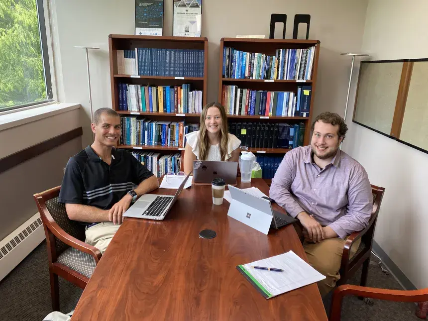 One female and two male students gathered around a meeting table with a bookcase behind.