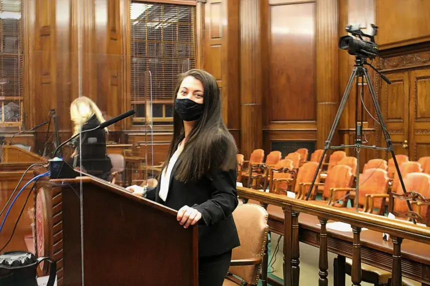 A person with a mask and long brown hair stands at the podium
