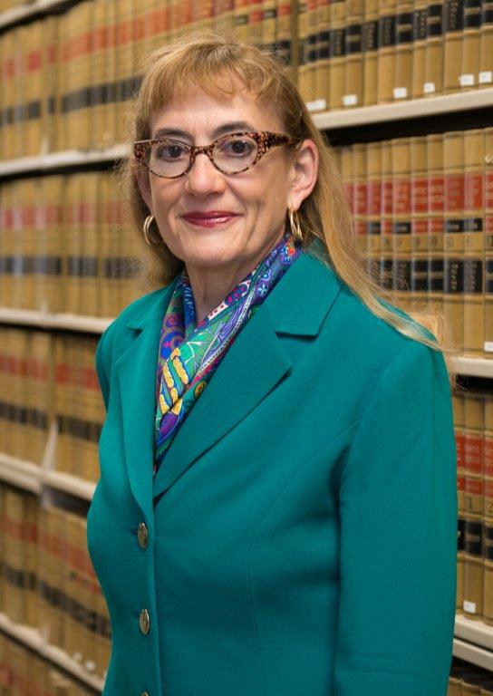 Woman with eyeglasses and long hair wearing a scarf and jacket against a row of books